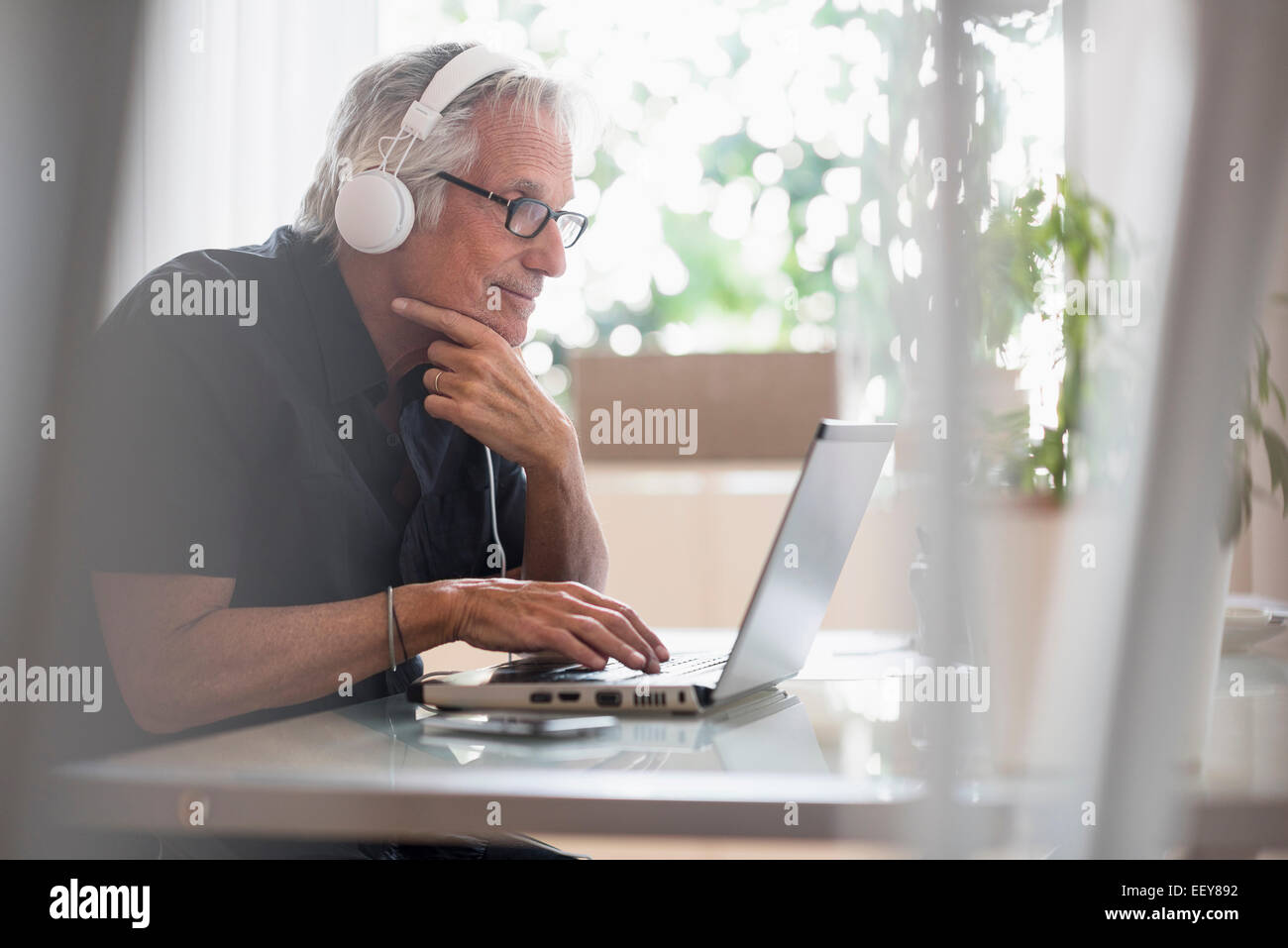 Senior uomo seduto in casa ufficio indossando le cuffie e utilizzando computer portatile Foto Stock