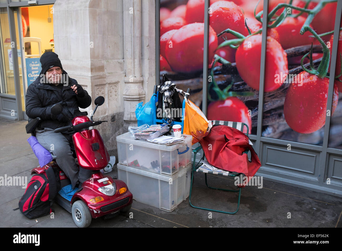 Un regolare appende fuori vicino al supermercato Tesco su Bishopsgate, London, Regno Unito. Questo uomo arriva sul suo scooter di mobilità per sedersi a chiacchierare con un grande problema venditore che funziona in questo passo di fronte alcuni grossi pomodori che sono parte di prodotti freschi di pubblicità per la catena di supermercati. Foto Stock