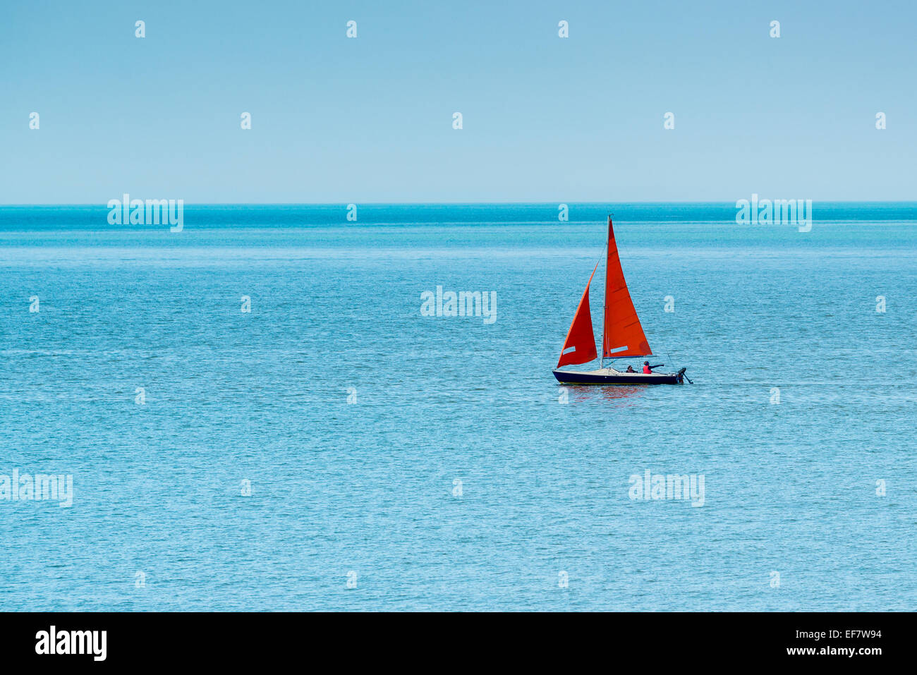 Unico dinghy a vela con il rosso-arancio di vele su una calma, azure, mare sotto un cielo blu chiaro. Foto Stock