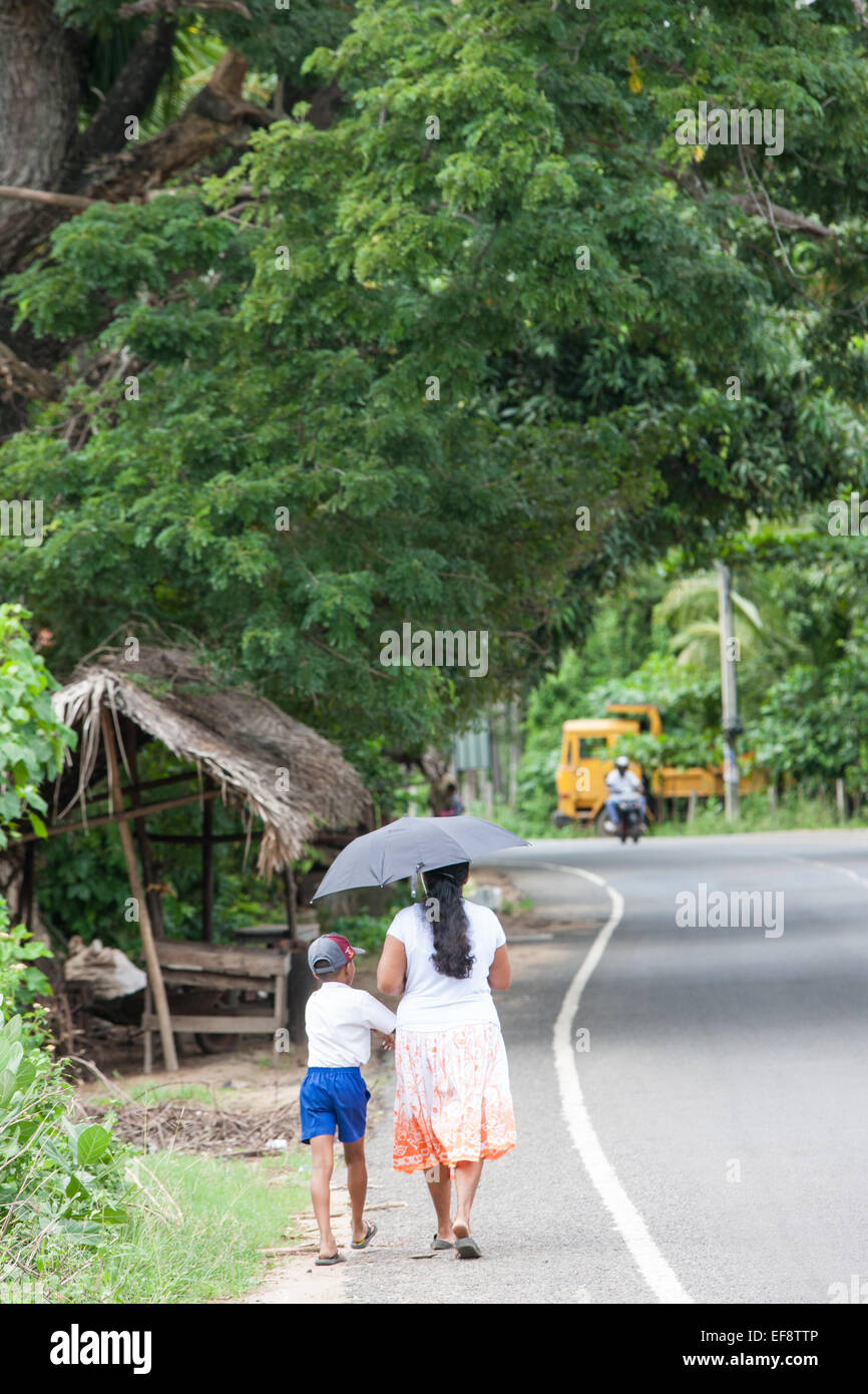 Famiglia locale a camminare a fianco di una strada rurale in una scuola gestita. Andare a scuola a piedi. A Tissa,Tissamaharama,Sri Lanka.Ombrello ombra.dello Sri Lanka, Foto Stock