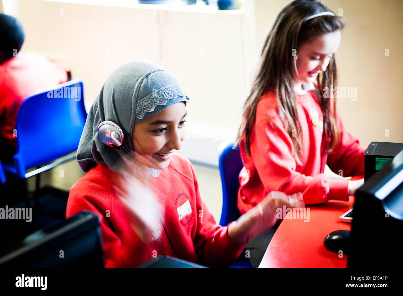 Ragazzina musulmana in velo usando le cuffie a un computer nella scuola primaria aula in tutto il Regno Unito Foto Stock