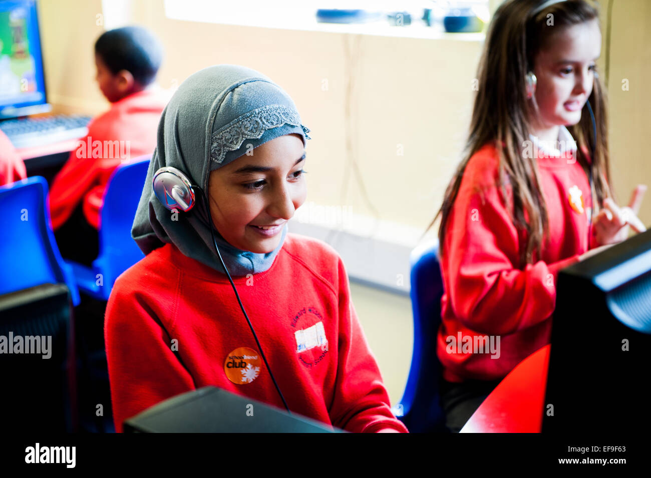 Ragazzina musulmana in velo usando le cuffie a un computer nella scuola primaria aula in tutto il Regno Unito Foto Stock