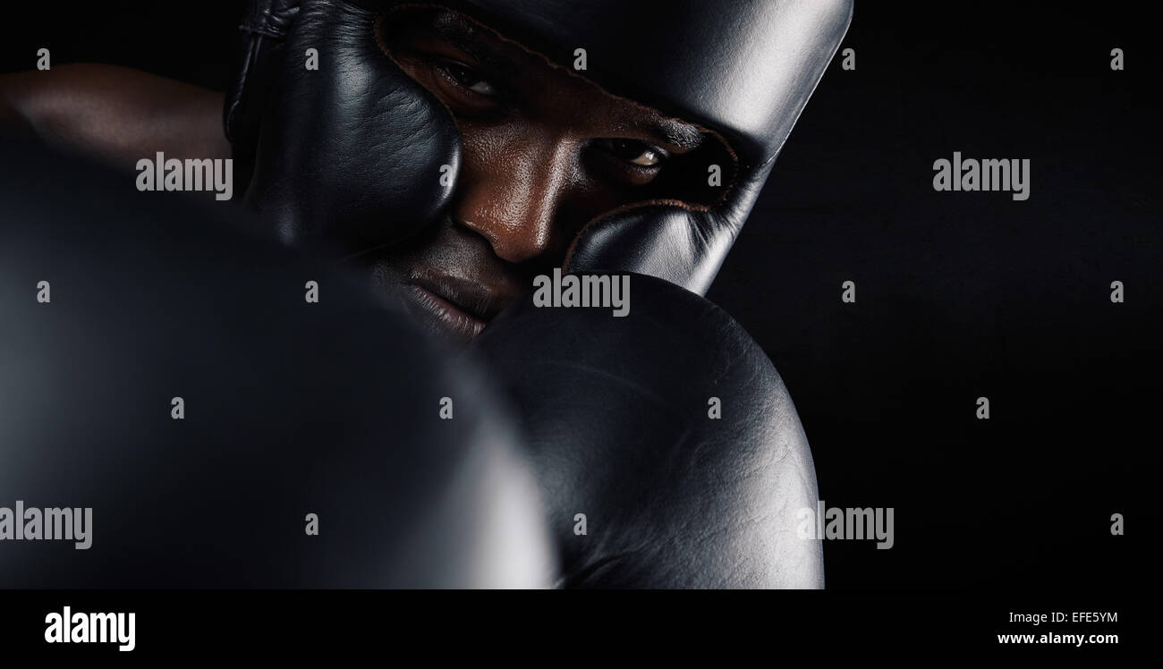 Close-up di immagine africana boxer maschio indossa testa protettiva guard e guanti di protezione contro lo sfondo nero. Giovane uomo che esercitano box Foto Stock