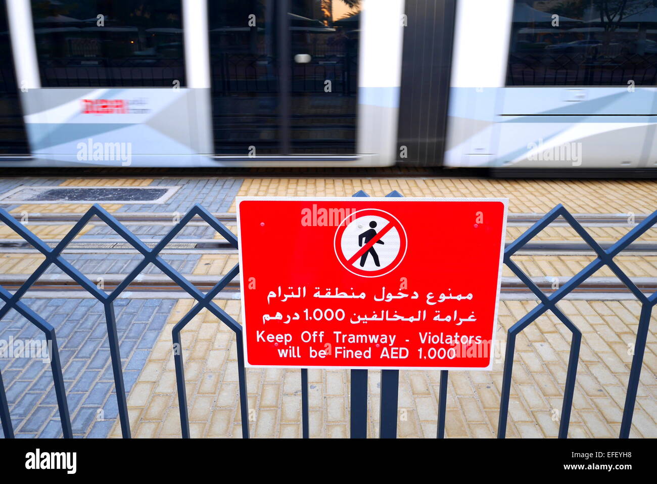 Un segno in arabo e in inglese avviso di persone di non camminare a piedi sui binari del tram, un tram in movimento dietro, Dubai, Emirati Arabi Uniti Foto Stock