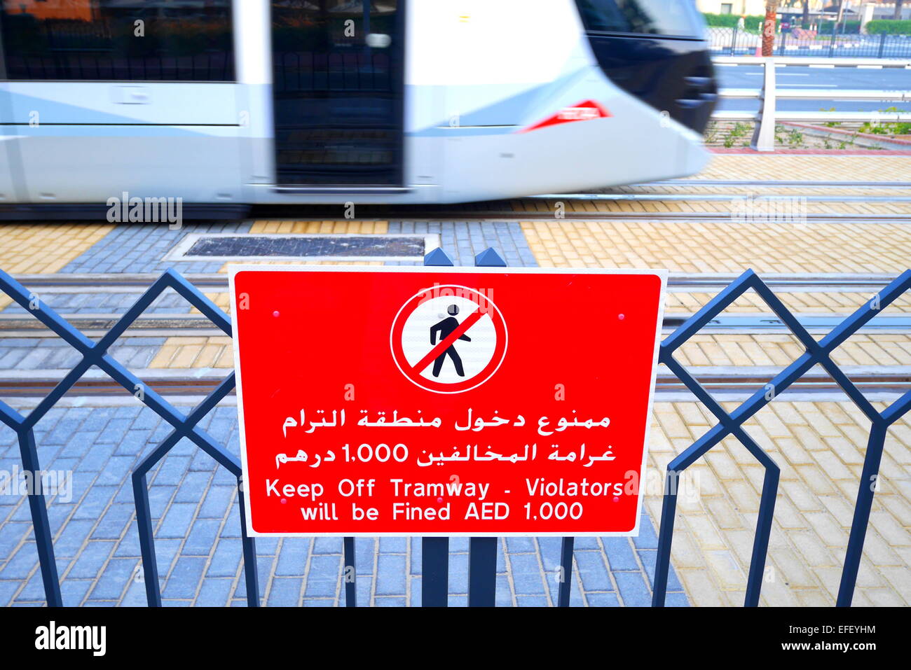 Un segno in arabo e in inglese avviso di persone di non camminare a piedi sui binari del tram, un tram in movimento dietro, Dubai, Emirati Arabi Uniti Foto Stock