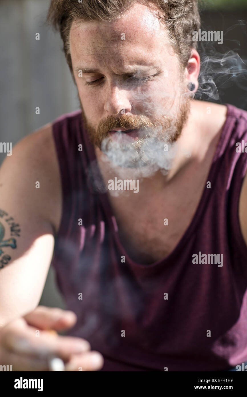 Un giovane uomo seduto al di fuori di una casa abbandonata e fumo di sigarette. Problemi di tossicodipendenza. Foto Stock