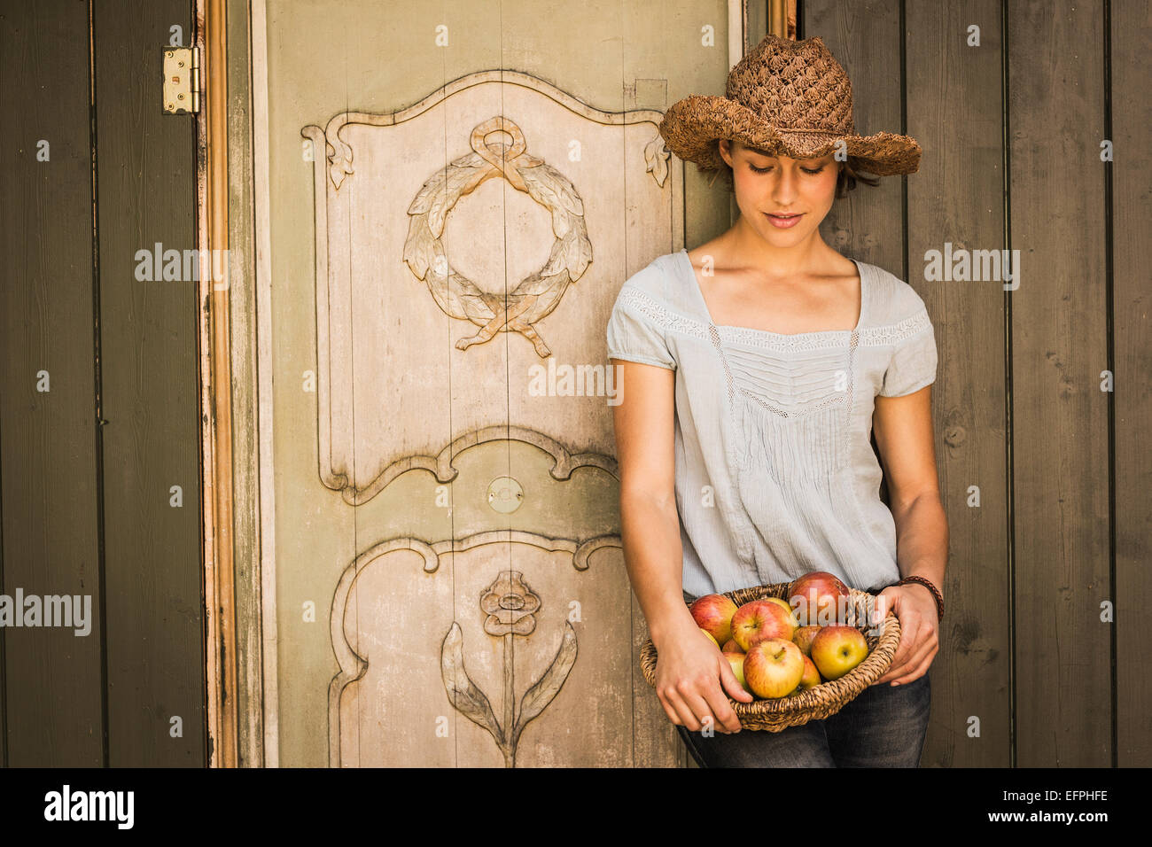 Giovane donna che indossa cappello di paglia, appoggiata contro un capannone, cestello di contenimento di mele Foto Stock