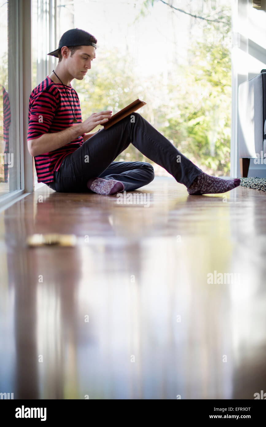 Uomo che indossa un cappello da baseball indietro, seduto sul pavimento in un salotto, guardando una tavoletta digitale. Foto Stock