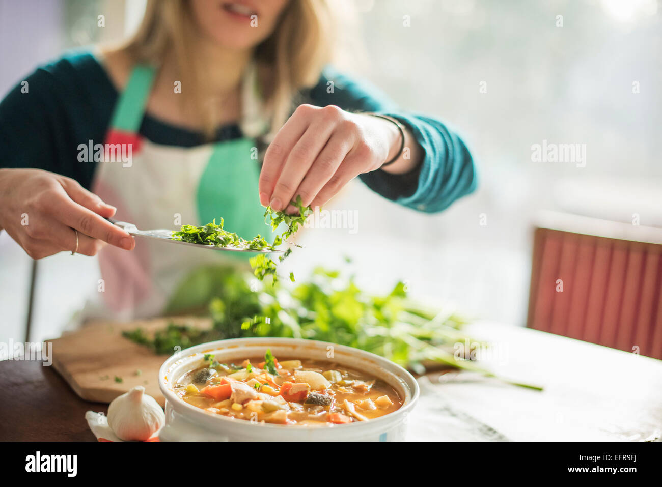 Una donna che indossa un grembiule, seduti a un tavolo, spargendo erbe in una ciotola di stufato di vegetali. Foto Stock