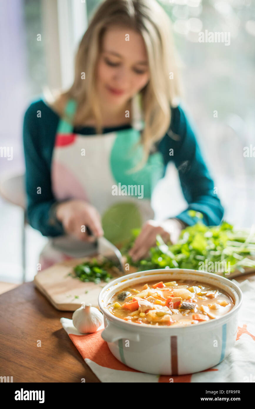 Una donna che indossa un grembiule, seduti a un tavolo, tritare erbe, una ciotola di stufato di vegetali in primo piano. Foto Stock