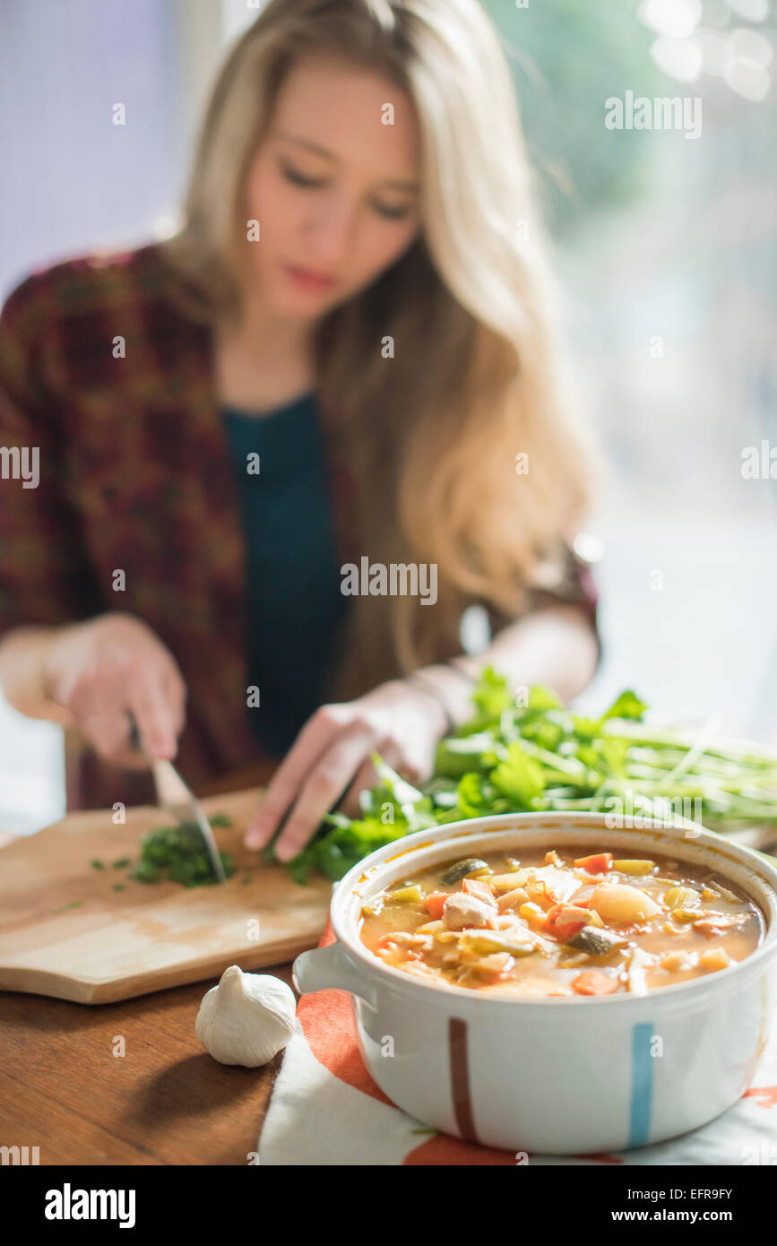Una donna seduta al tavolo, tritare erbe, una ciotola di stufato di vegetali in primo piano. Foto Stock