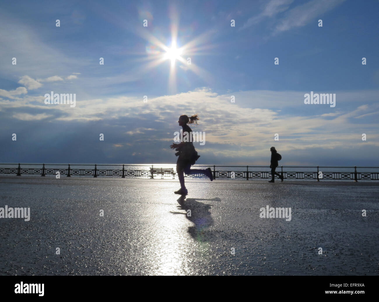 Una ragazza corre pareggiatore lungo prati di Hove promenade casting la sua ombra nella luce del sole della mattina accanto alla luccicante mare Foto Stock