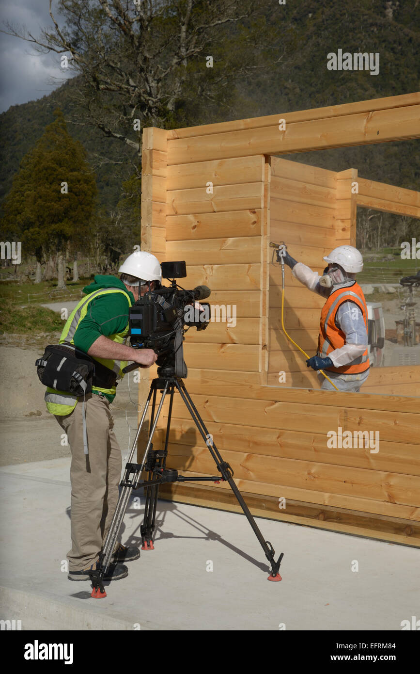 Un documentario film maker un commerciante verniciatura a spruzzo la parete del legno di un edificio industriale con il preservante del legno Foto Stock