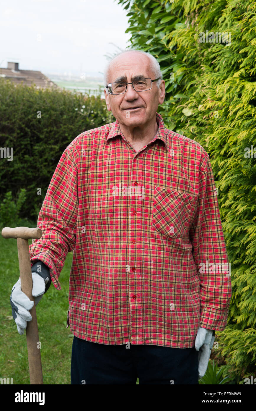 Uomo anziano in piedi in giardino, indossare abiti da lavoro e azienda vanga Foto Stock