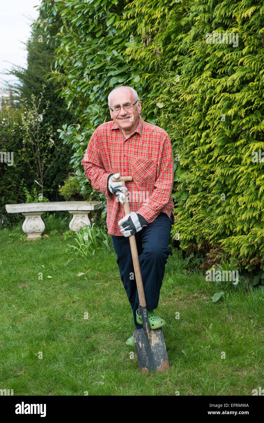 Uomo anziano in piedi in giardino, indossare abiti da lavoro e azienda vanga, felice Foto Stock