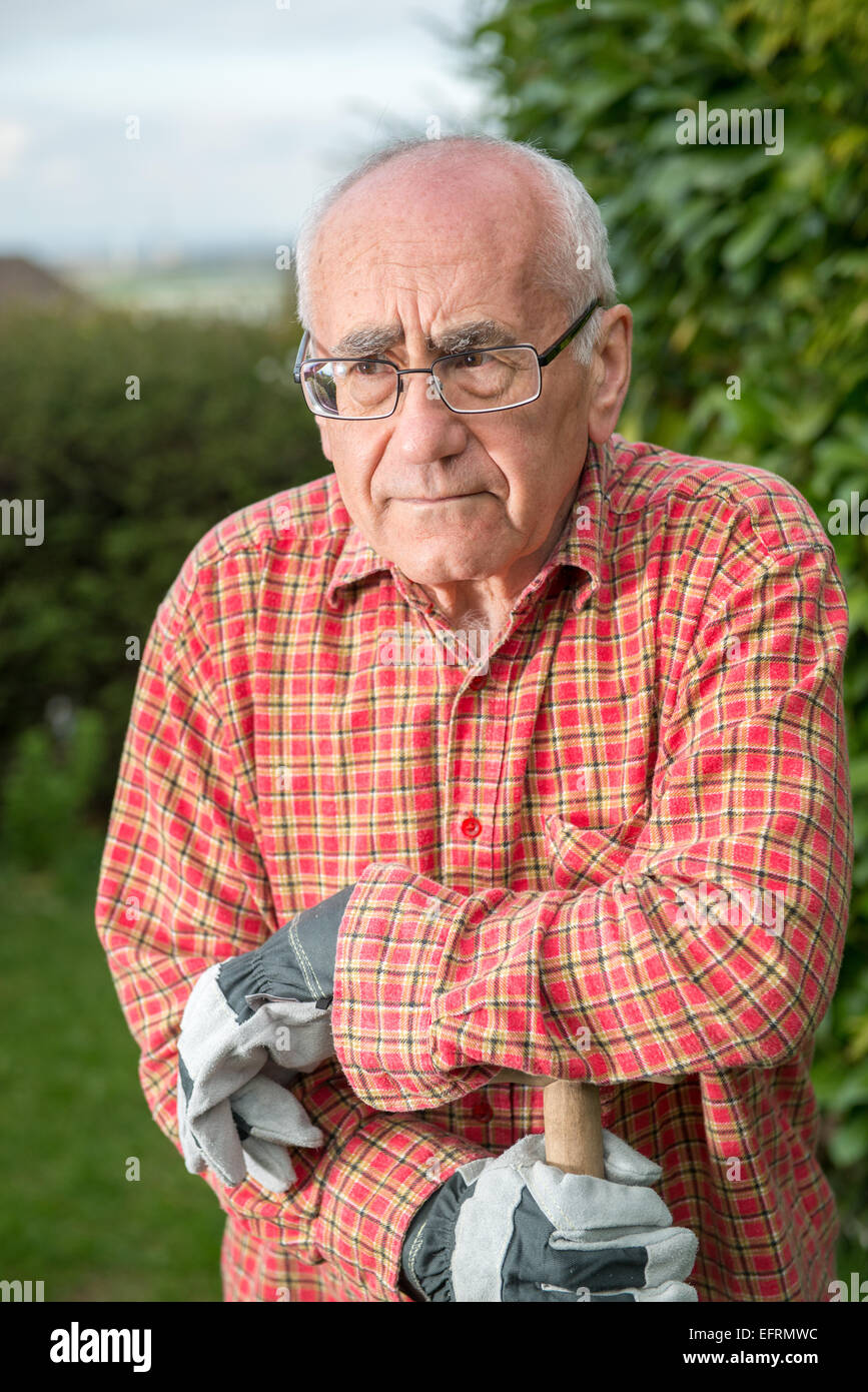 Uomo anziano in piedi in giardino, indossare abiti da lavoro e azienda vanga Foto Stock