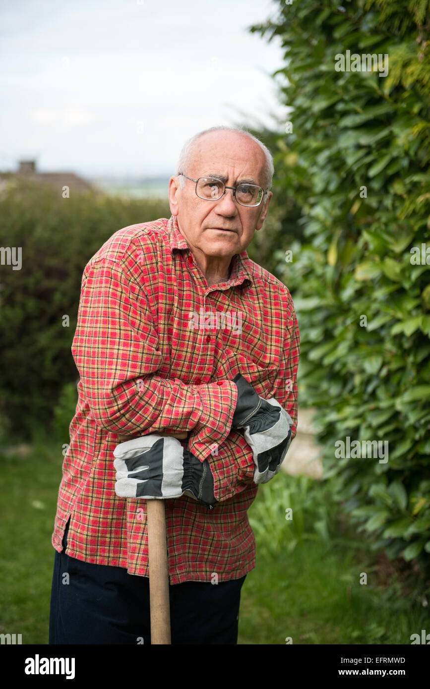 Uomo anziano in piedi in giardino, indossare abiti da lavoro e azienda vanga, faccia woried Foto Stock