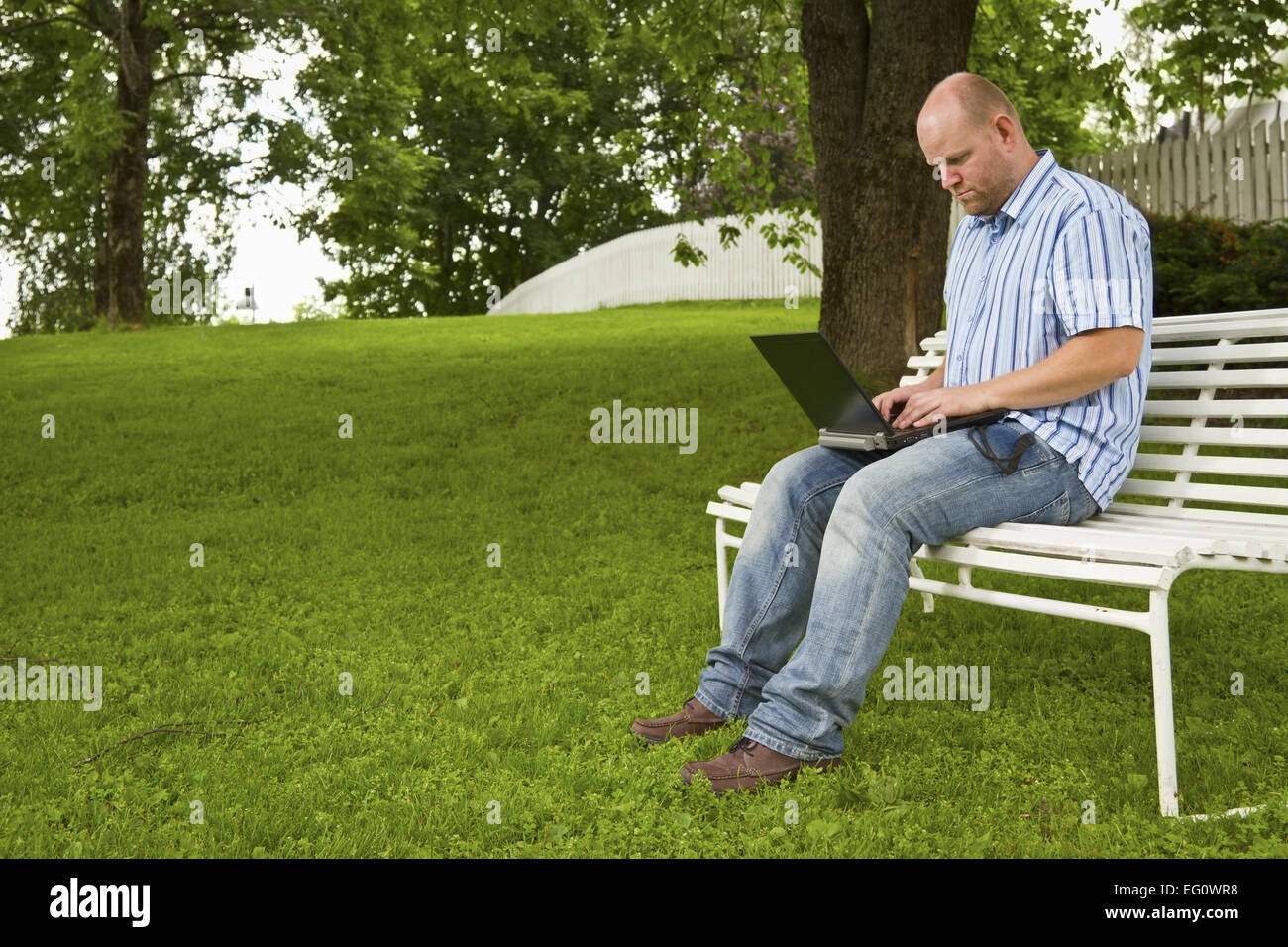 Un concentrato di lavoro uomo / studiando un libro in un parco. Seduta su una panchina con un libro e un computer portatile. Foto Stock