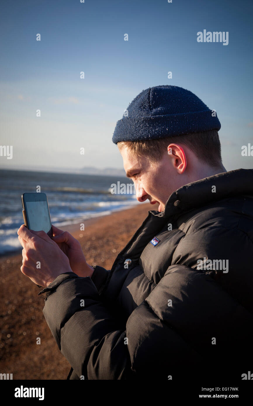 Giovane uomo che indossa 'puffa' giacca e cappello di lana in inverno, scatta una fotografia con un ipad su una spiaggia di ciottoli. Foto Stock