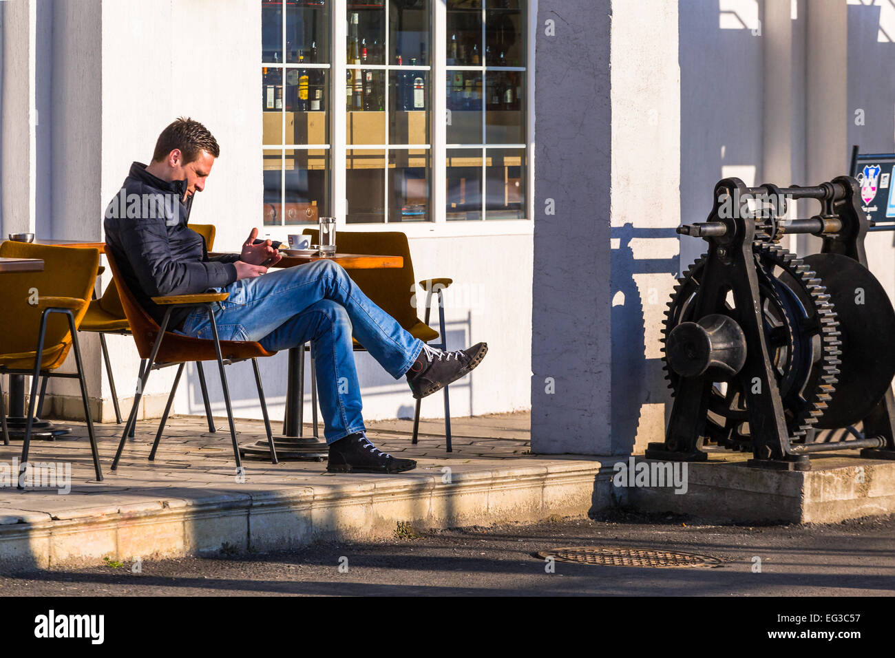 Un uomo è seduto fuori da un bar e controlla il suo telefono in Savamala Belgrado. bottiglie di alcolici può essere visto nella finestra Foto Stock