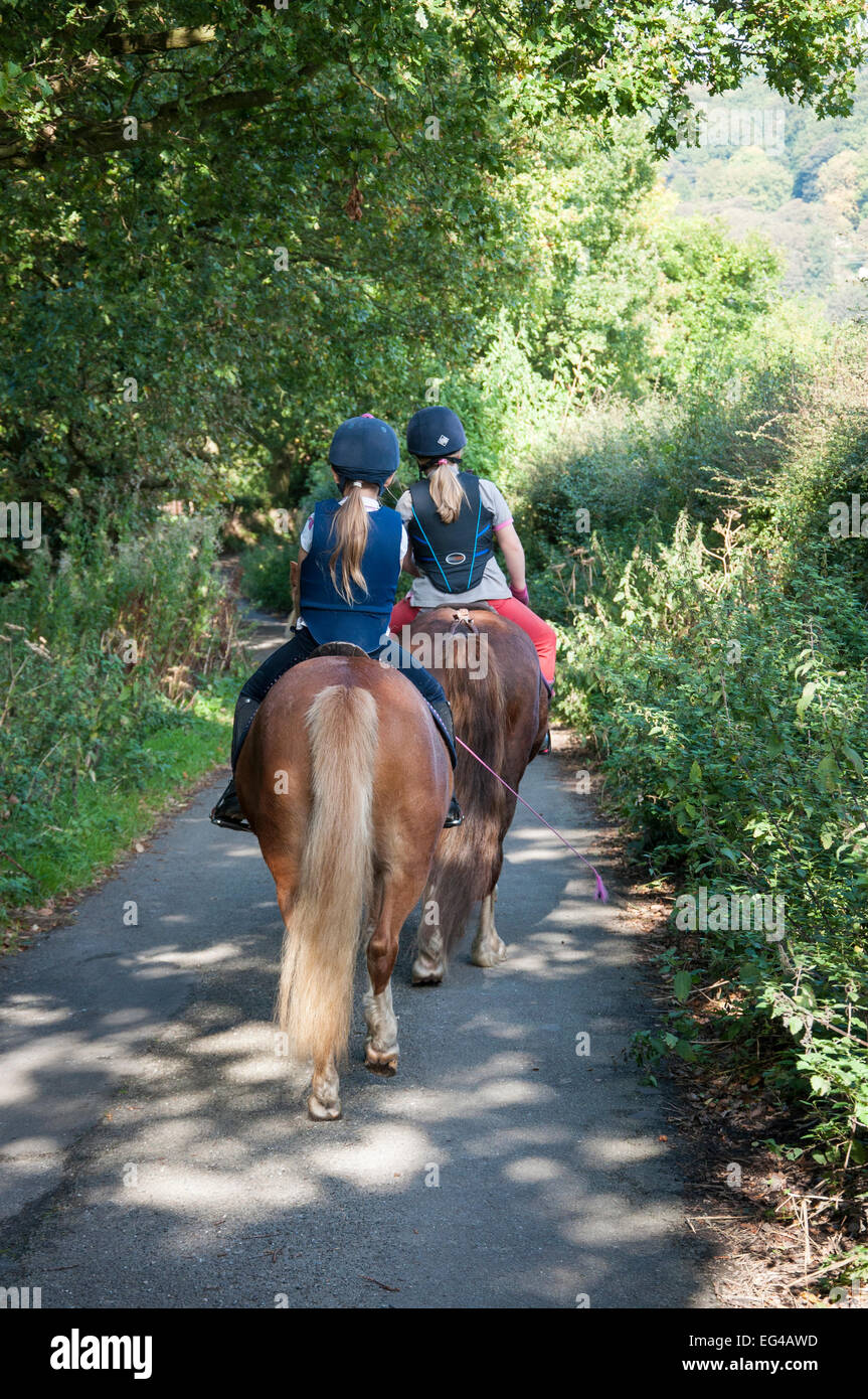 Due giovani ragazze cavalcare i pony in un paese di lingua inglese lane in tarda estate sunshine. Foto Stock