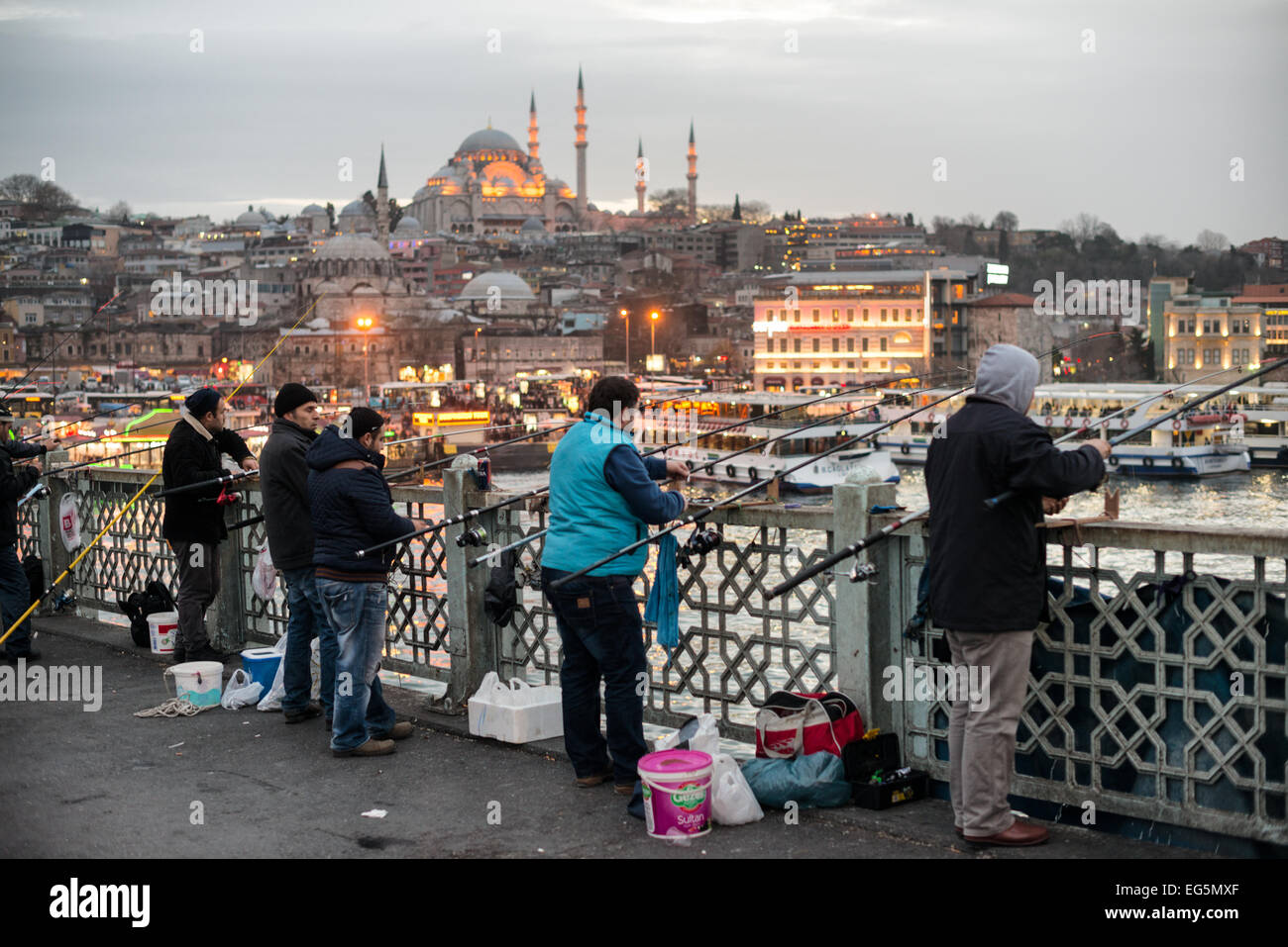 ISTANBUL, Turchia / Türkiye - i pescatori fiancheggiano il bordo del ponte di Galata con le loro linee sul lato del Corno d'Oro. Attraversando il Corno d'Oro e collegando Eminonu con Karakoy, il ponte Galata è un ponte a due livelli che gestisce il traffico stradale, del tram e pedonale al piano superiore con ristoranti e bar al livello sottostante. In lontananza c'è la Moschea Suleymaniye. Foto Stock