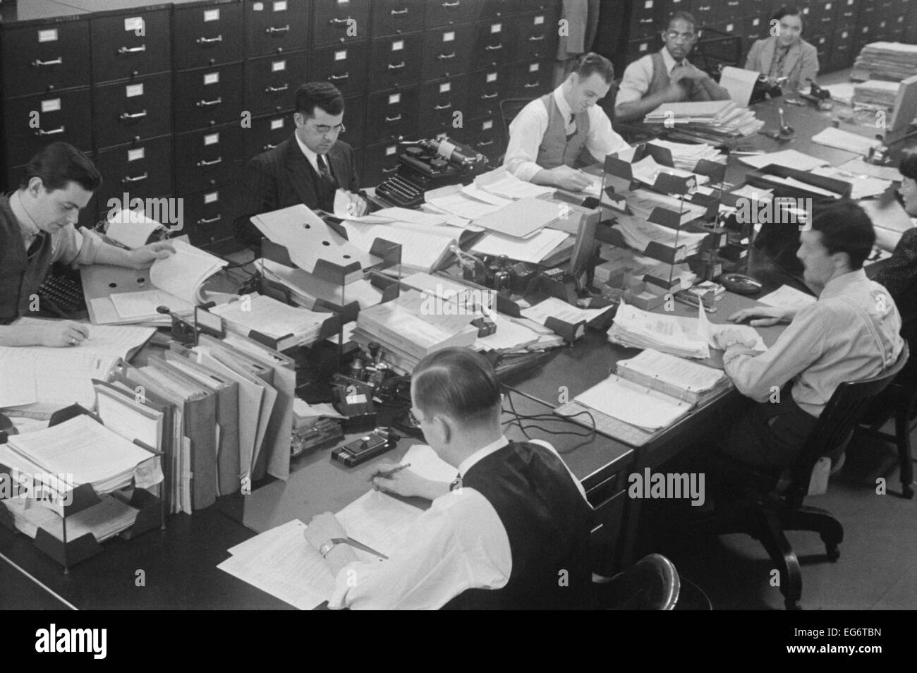 Sei uomini e due donne al lavoro in un governo degli Stati Uniti in ufficio. Essi condividono un lungo tavolo, coperto con documenti, notebook e Foto Stock