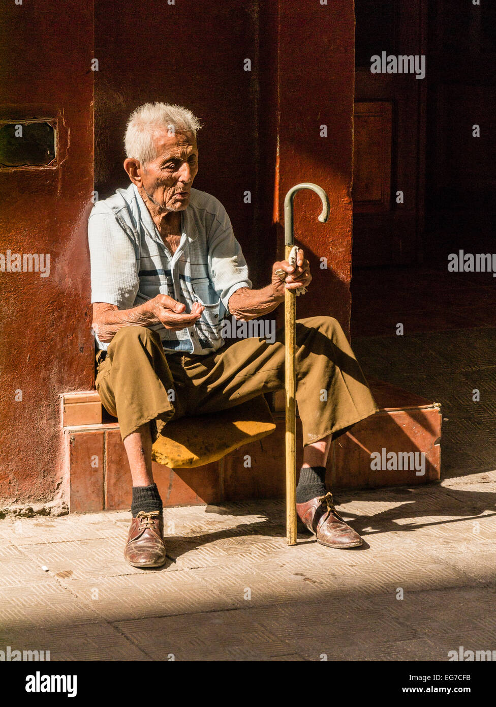 Un anziano maschio mendicante cubano seduto su una porta stoop tenendo la sua mano per denaro con una canna da zucchero a l'Avana, Cuba. Foto Stock