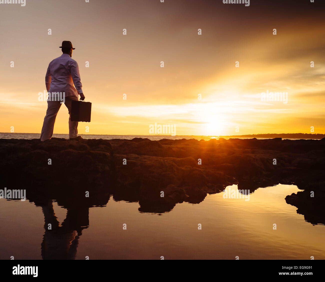 Uomo che cammina sulla spiaggia al tramonto portando con sé una valigia Foto Stock