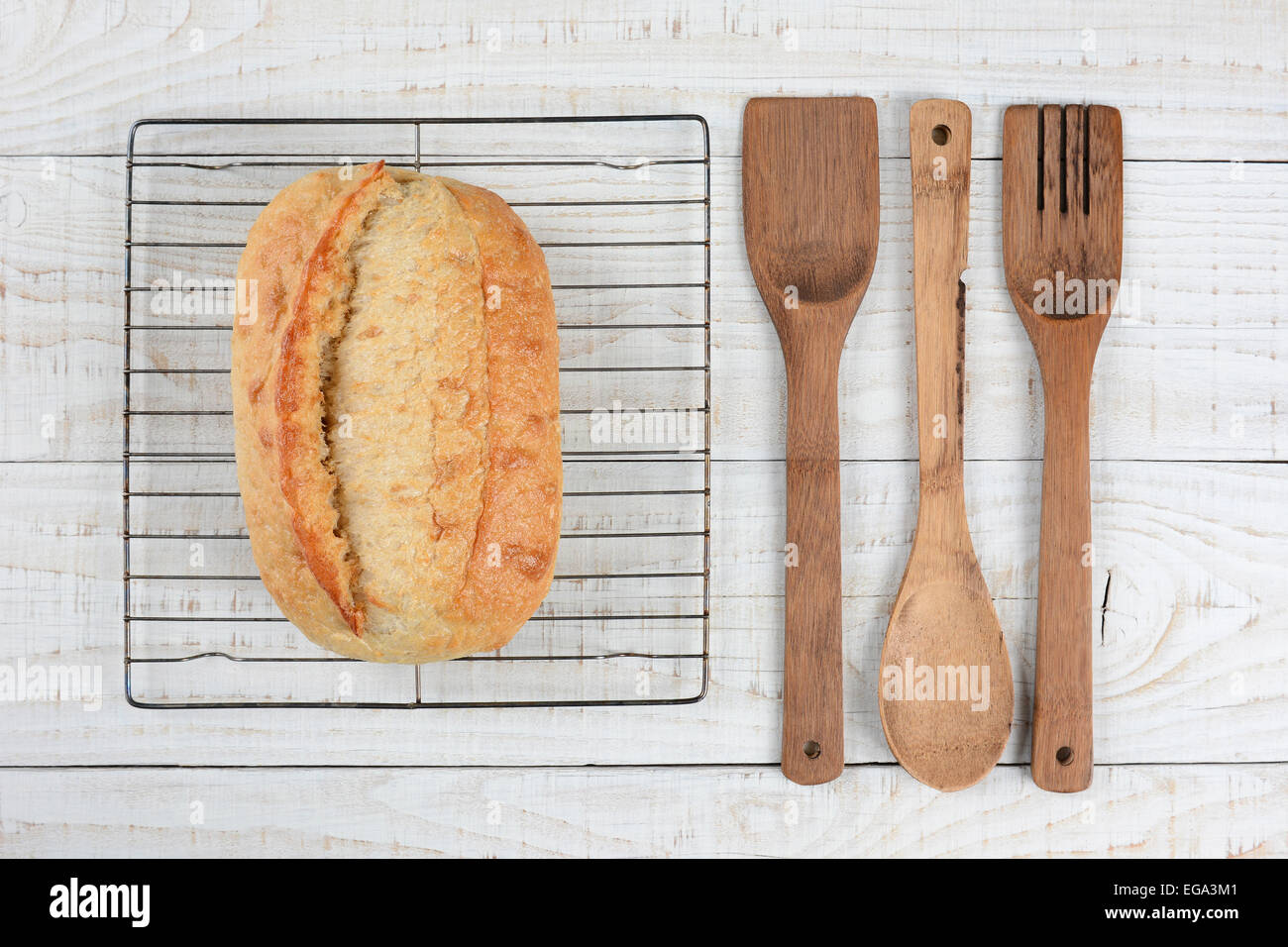 Alta angolazione di una fresca di forno pagnotta di pane su una griglia di raffreddamento con il cucchiaio di legno, una spatola e la forcella. Formato orizzontale Foto Stock