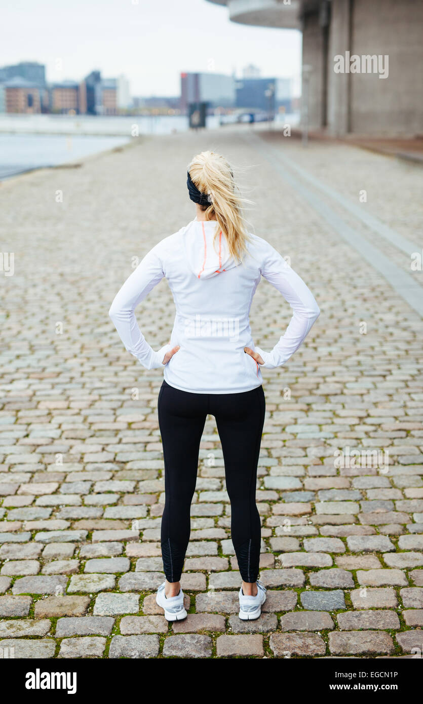 Vista posteriore della giovane donna con le mani sui fianchi in piedi sul marciapiede. Modello di fitness di guardare avanti per una città esegui. Foto Stock