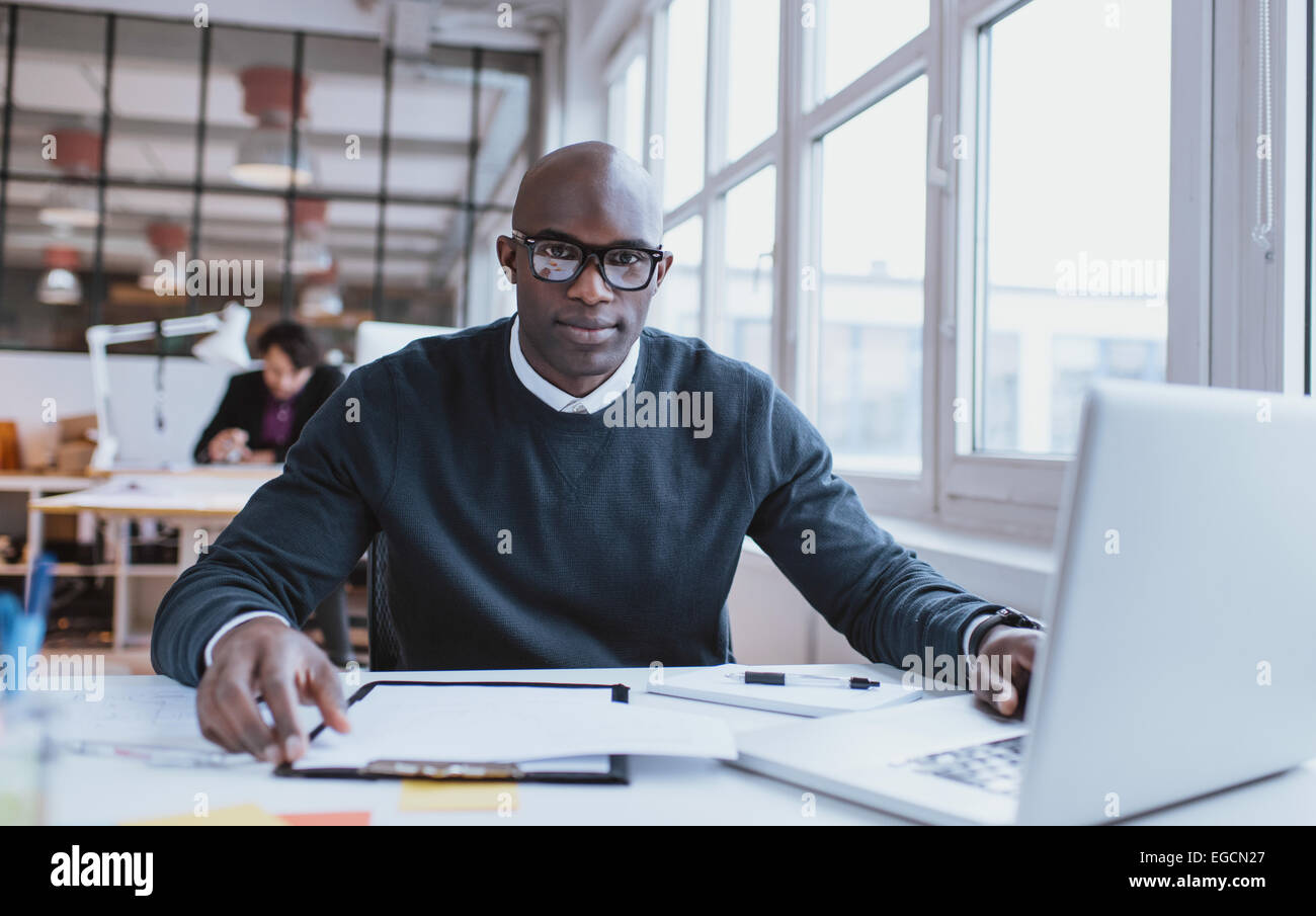 Ritratto di bello giovane africano alla sua scrivania con il computer portatile. L'uomo africano guardando la fotocamera mentre si è al lavoro. Foto Stock