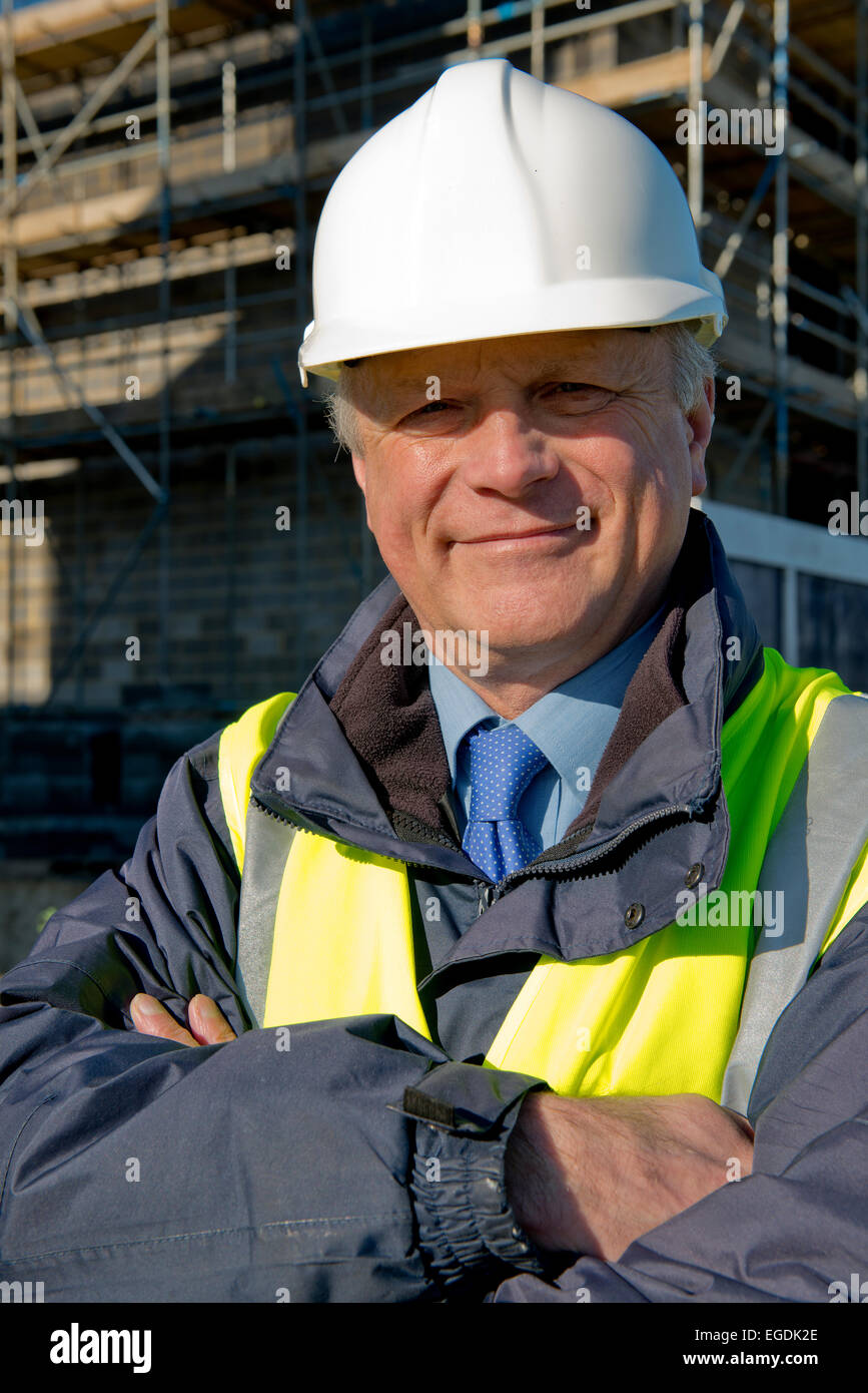 Sorridente lavoratore edile con le braccia piegate, in piedi di fronte ad un edificio sito. Foto Stock