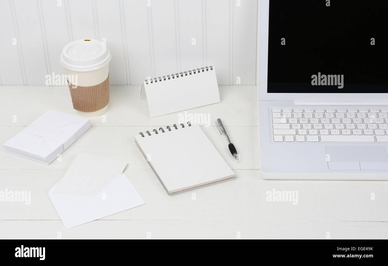 Primo piano di un bianco puro scrivania con computer portatile, monouso tazza da caffè, appunti, buste e penna. Una scrivania in legno con una parete beadboard Foto Stock