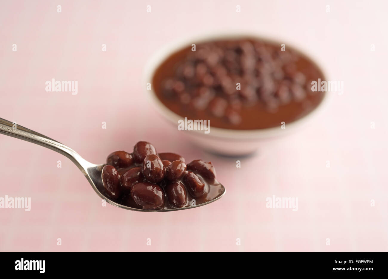 Un cucchiaio in primo piano riempito con i fagioli neri in una salsa chili con una ciotola in background su una scacchiera rosa tabella cl Foto Stock