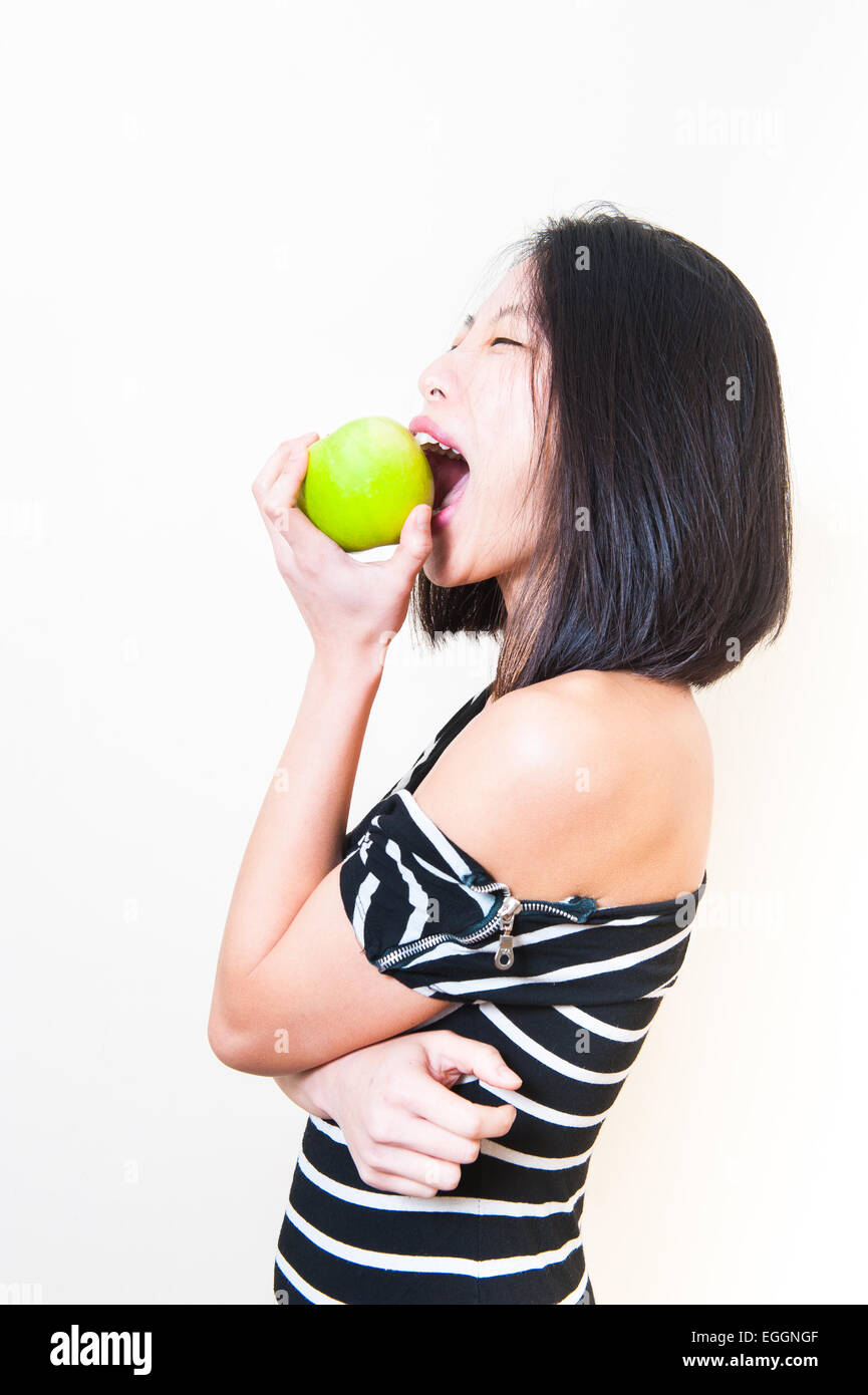 Giovane bella donna asiatica di mordere mela verde con espressione arrabbiato, profilo su sfondo bianco Foto Stock