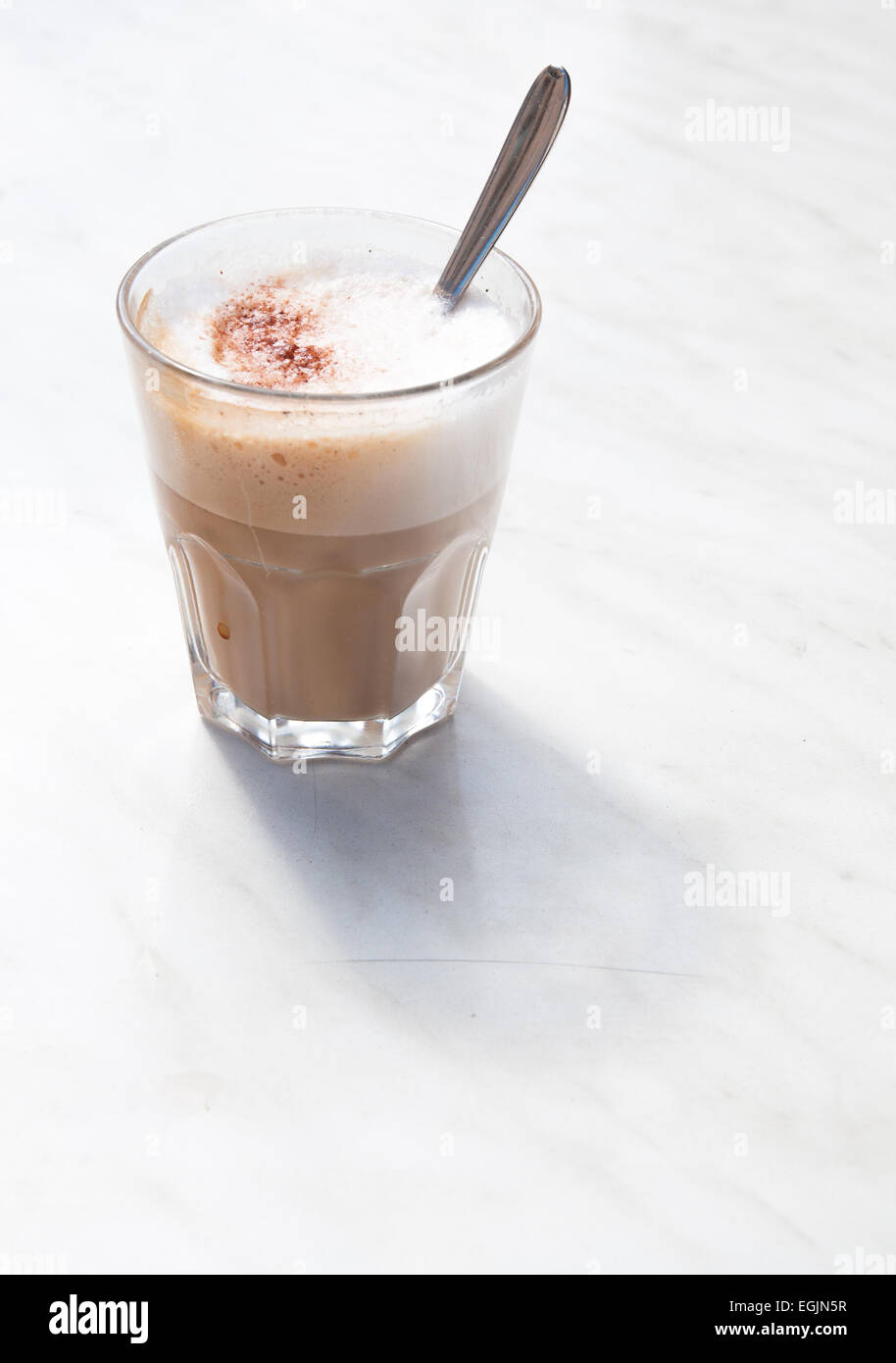 Cappuccino schiumoso in un caffè di vetro con cucchiaio su marmo bianco tabella all'aperto, dal lato. Foto Stock