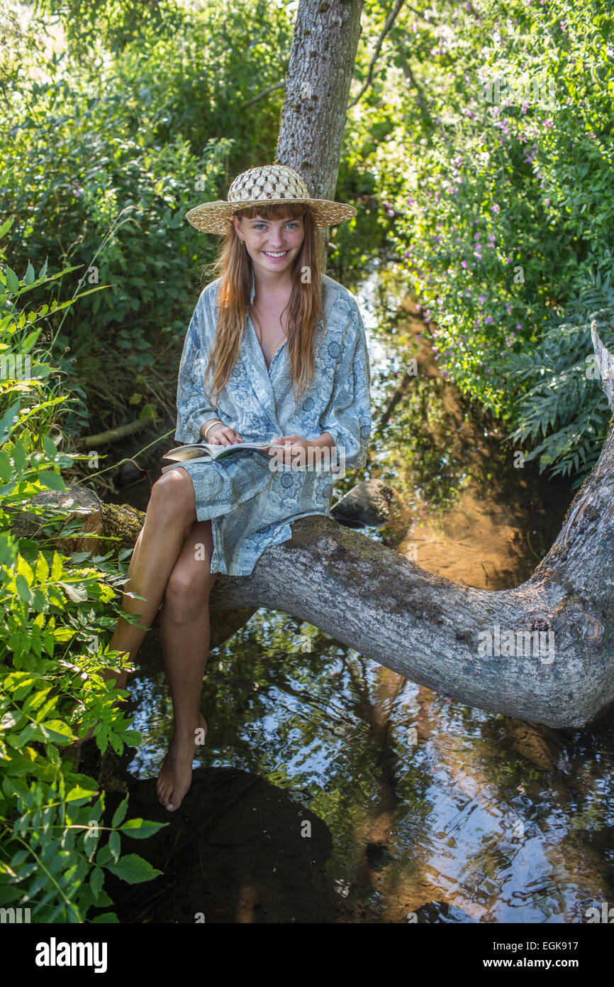 Ragazza con un cappello a sorridere al visualizzatore mentre è seduto in un albero più di una certa quantità di acqua e la lettura di un libro al di fuori nella luce del pomeriggio. Foto Stock