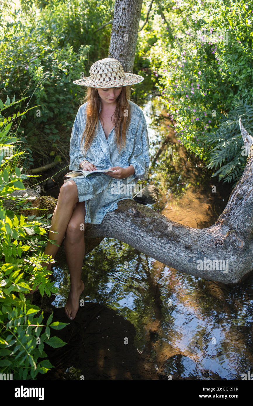 Ragazza con un cappello seduti in un albero più di una certa quantità di acqua e la lettura di un libro al di fuori nella luce del pomeriggio. Foto Stock