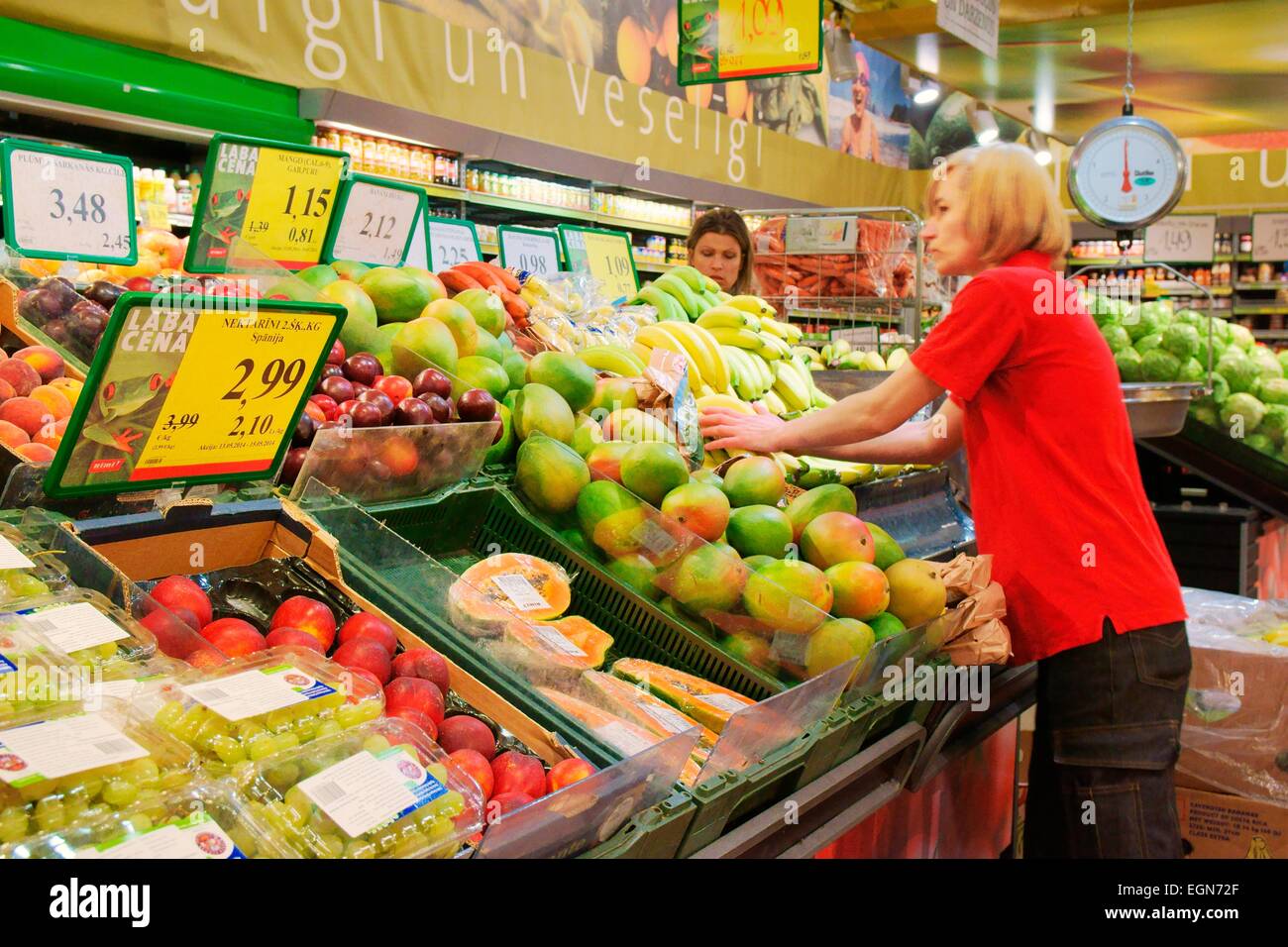 Riga, Lettonia. Interno del ipermercato Rimi lettone store. Assistente di ripiani di impilamento al di frutta fresca e vegetali di stallo del contatore Foto Stock