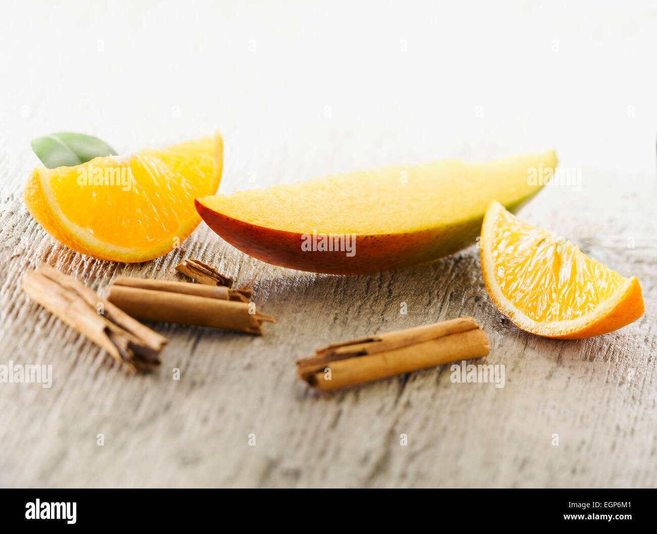 Mango, Mangifera indica. Uno strato disposto con quarti di Orange e pezzi rotti di stecca di cannella sul pallido, angosciato, sullo sfondo di legno. Messa a fuoco selettiva. Foto Stock