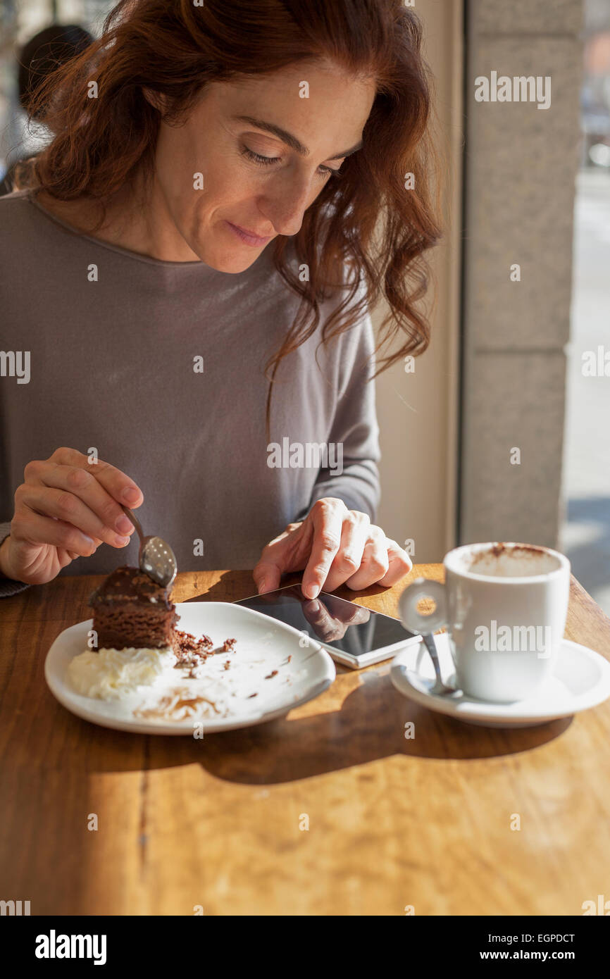 Donna prendendo la torta al cioccolato pezzo con cucchiaio accanto al bianco piccola tazza cappuccino e caffè toccando Telefono cellulare schermo bianco su Foto Stock