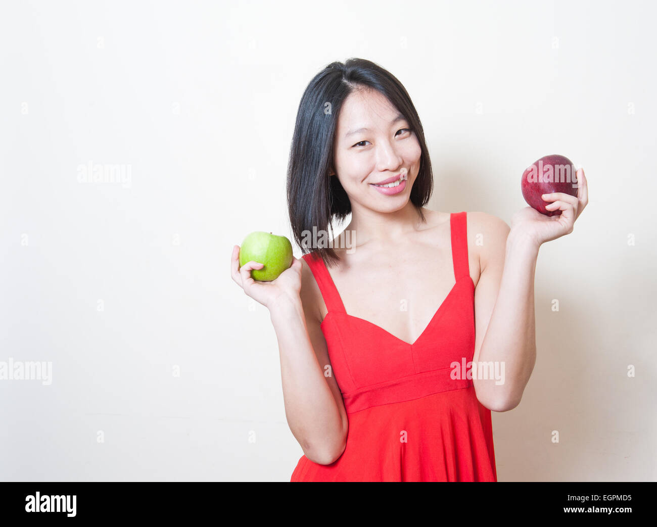 Giovane bella donna asiatica in abito rosso sorridente e mostrando il verde e le mele rosse sulle sue mani su sfondo bianco Foto Stock