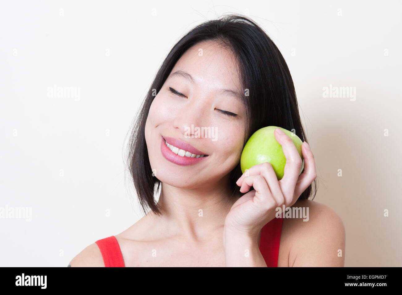 Giovane bella donna asiatica in abito rosso closeup sorridente con il chiudere gli occhi, mela verde sulla sua mano vicino a faccia su sfondo bianco Foto Stock
