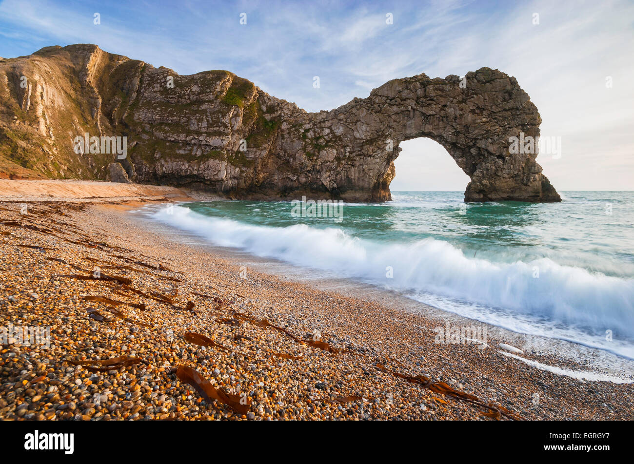 Un'onda si rompe sulla spiaggia di ciottoli con il famoso arco naturale di Durdle porta dietro. Foto Stock