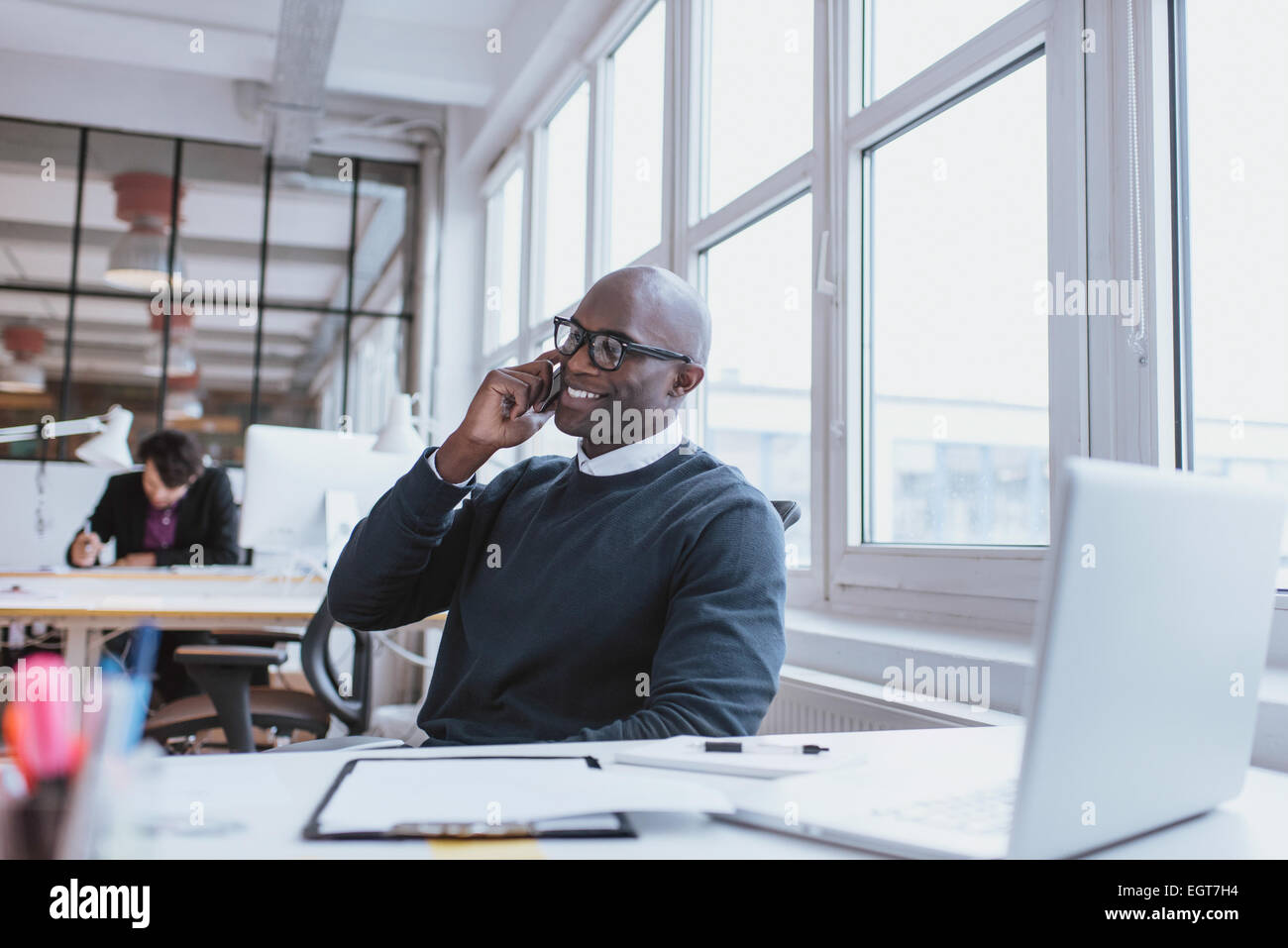 Giovane uomo che parla per il suo telefono cellulare in ufficio. African executive seduto alla sua scrivania con computer portatile Foto Stock