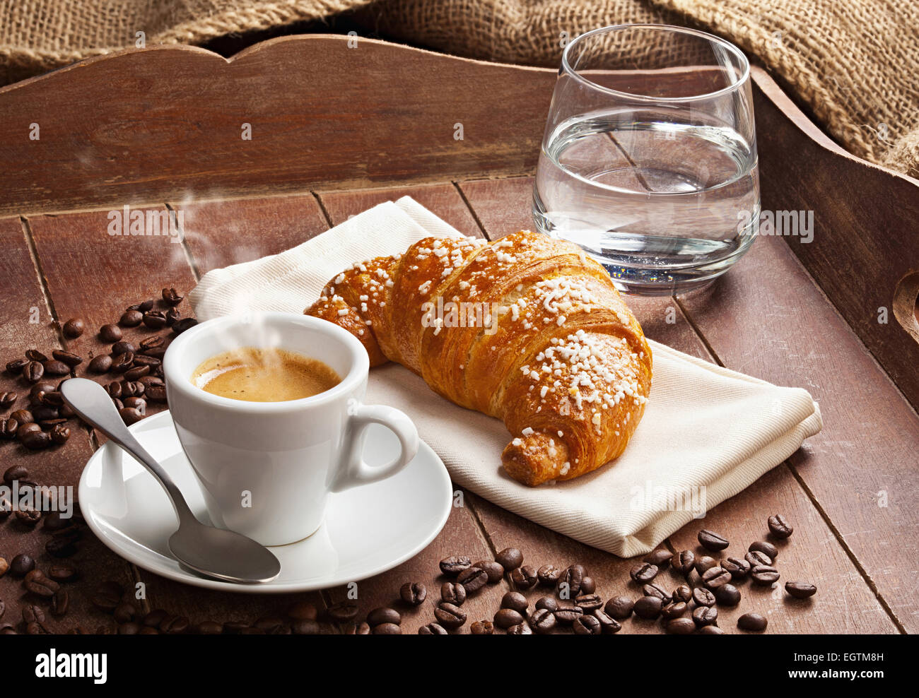 Espresso con croissant e un bicchiere di acqua nella vaschetta di legno marrone. Foto Stock