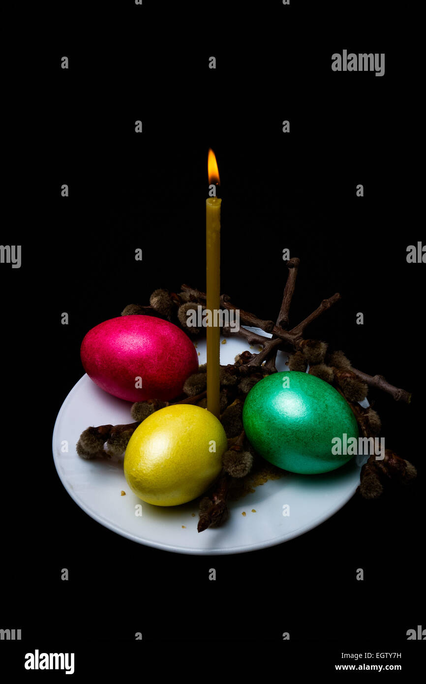 La masterizzazione di candele di cera, tre uova di Pasqua di rosso, giallo e verde, rametti di salice con boccioli soffici sul piattino Foto Stock
