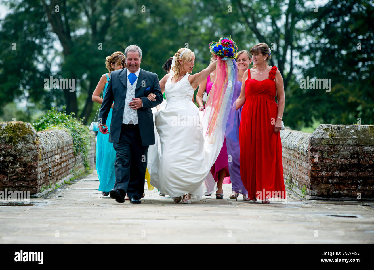 Una sposa danze eccitato come lei cammina verso la sua cerimonia di nozze con il padre della sposa e le sue damigelle hanno. Foto Stock