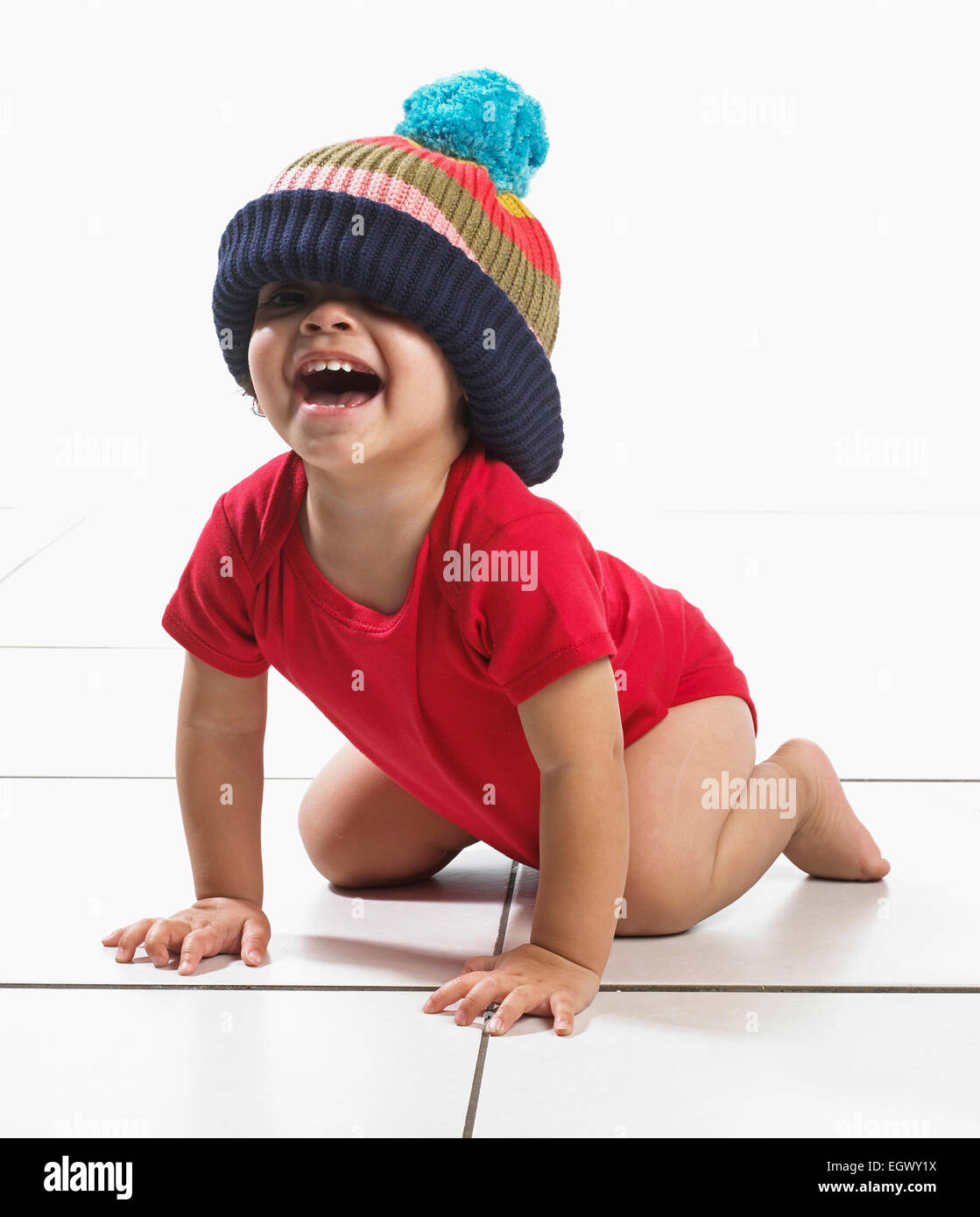 Ragazzo (16 mesi) inginocchiati sul pavimento ridendo, indossando il giubbotto e maglia di grandi dimensioni bobble hat Foto Stock